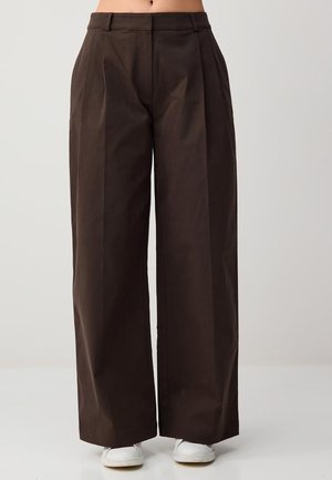 Brązowe spodnie damskie z wysokim stanem i szerokimi nogawkami, noszone z białymi trampkami, widoczne od talii do podłogi na jednolitym tle.