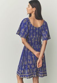 Robe bleue avec de courtes manches volantées, ornée de motifs en feuilles d'or, taille smockée et ourlet agrémenté d'accents en étoiles dorées. Tissu texturé.