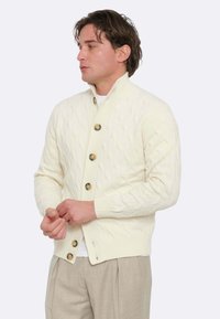 Cardigan crema a maglia cable con colletto alto, quattro grandi bottoni tondi, polsini a coste e design aderente. Indossato sopra una camicia bianca.