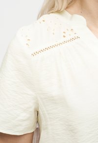 Γυναίκα που φοράει μπλούζα σε κρεμ χρώμα με κοντά μανίκια και δαντελωτές λεπτομέρειες κοντά στον ώμο και το γιακά.