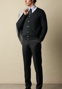 Dunkelgraue Strickjacke mit V-Ausschnitt und knöpfbarem Vorderteil, getragen über einem hellblauen gestreiften Hemd und einer dunklen gepunkteten Krawatte; schwarze Hose.