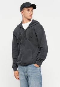 Young man wearing a black cap, charcoal gray zip-up hoodie with front pockets, and light blue jeans, looking to the side against a plain background.
