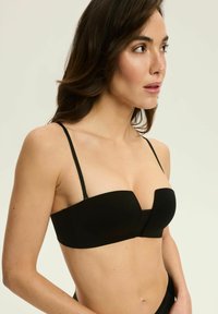 Czarny strapless stanik z cienkimi regulowanymi ramiączkami, gładkiego materiału, o konstrukcji z delikatnym szwem w kształcie litery V na środku.