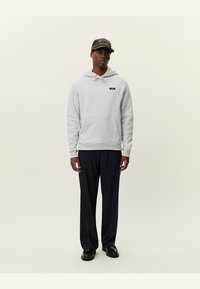 Sudadera con capucha gris claro, con bolsillo frontal y parche de logo negro; combinada con pantalones oscuros de color azul marino y una gorra oscura con texto bordado.