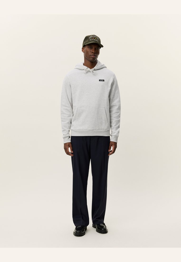 Sudadera con capucha gris claro, con bolsillo frontal y parche de logo negro; combinada con pantalones oscuros de color azul marino y una gorra oscura con texto bordado.
