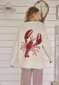 Bijeli široki džemper s motivom crvenog jastoga na leđima, izrađen od teksturirane pletene tkanine s plavim obrubom na rubu i manžetama.