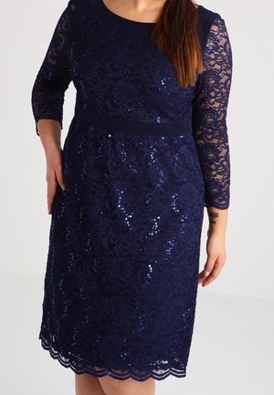 Kobieta w granatowej koronkowej sukience z rękawami do łokci i subtelnymi cekinami, stojąca z jedną ręką na biodrze.