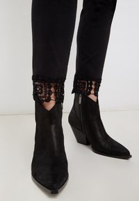 Černé semišové kotníkové boty s špičatou špičkou a bočním zipem, kombinované s černými kalhotami se zdobenými krajkovými manžetami.