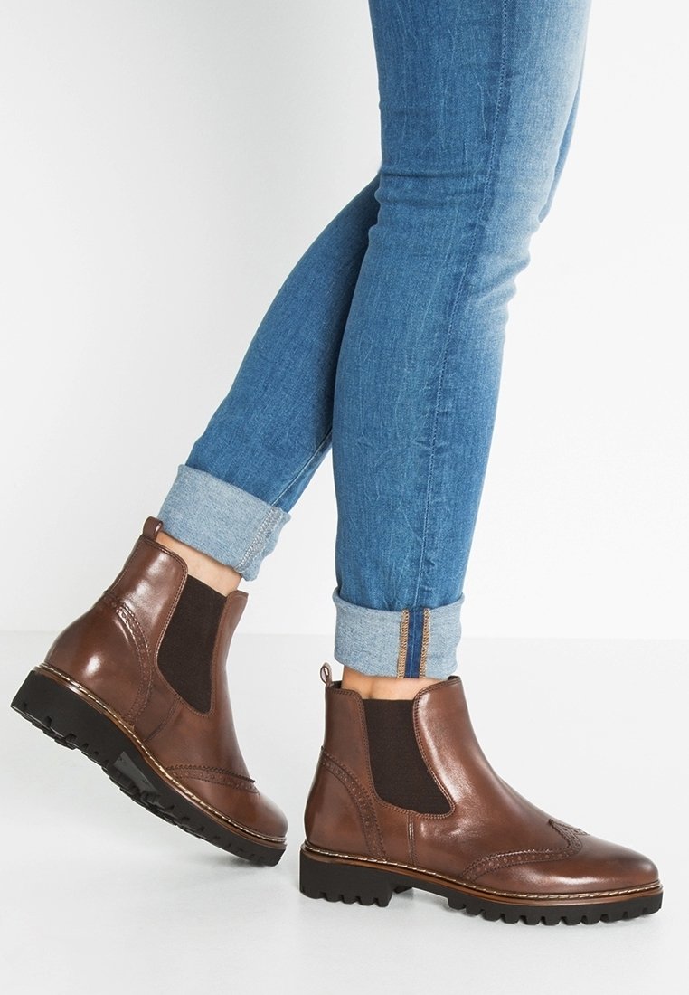 Bottines en cuir marron avec des panneaux élastiques sur les côtés, un design cousu et une semelle robuste, associées à un jean bleu retroussé.