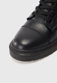 Zapatilla negra de cuero con punta redonda, parte superior texturizada y cordones negros, con una suela de goma gruesa y detalles de costura a lo largo de los bordes.