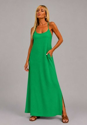 Femme aux longs cheveux blonds portant une robe maxi verte sans manches avec une fente latérale et des sandales marron, posant avec une main dans la poche.