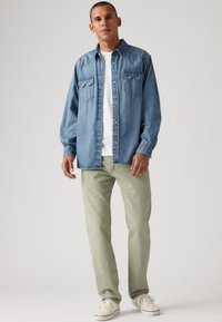 Helle blaue Denim-Hemd mit zwei Brusttaschen und silbernen Knöpfen, über einem weißen T-Shirt getragen, kombiniert mit hellgrünen Hosen und beigen Schuhen.
