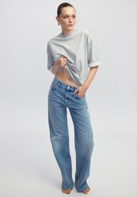Grijze, T-shirt met korte mouwen en een witte manchette, gecombineerd met hooglopende, lichtblauwe wijde jeans. Model poseert blootsvoets en toont de outfit.