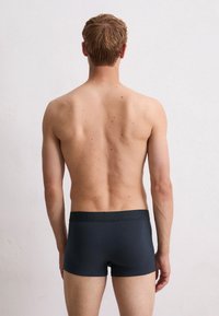 Giovane uomo con capelli castano chiaro in piedi di fronte a un muro bianco semplice, indossando boxer neri, mostrando la schiena e le braccia scoperte.