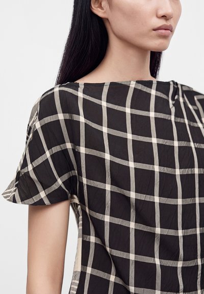 Femme portant un chemisier à manches courtes à carreaux noirs et beiges, avec un col rond et une texture de tissu lisse.