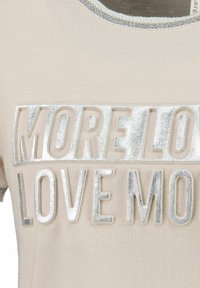 Světle béžové tričko s kulatým výstřihem, na kterém je nápis "MORE LOVE" a "LOVE MORE" z vypouklých metalických stříbrných písmen. Měkký textilní povrch.