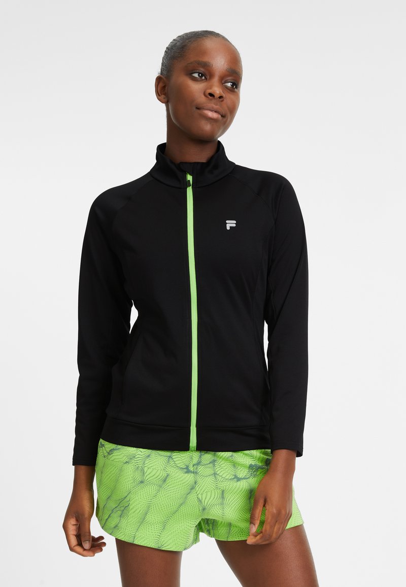 Giacca sportiva nera con colletto alto e cerniera verde. Abbinata a pantaloni corti verdi vivaci con un motivo testurizzato.