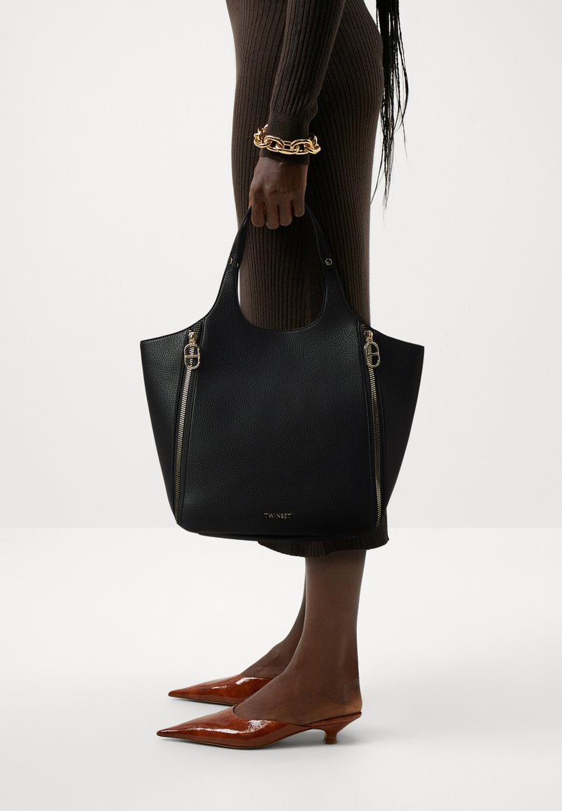 Zwarte leren tote bag met twee gouden ritsaccenten, met een afgeronde bovenkant en brede opening. Getoond met spitse bruine hakken.