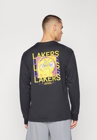 Juodas ilgomis rankovėmis marškinėliai su ryškiai geltona ir violetine grafika, vaizduojančia krepšinį, ir užrašų „Lakers“ ant nugaros. Minkšta medvilnė.