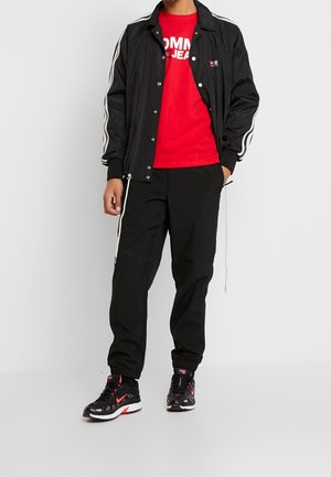 Sort vindjakke med hvide striber, rød t-shirt med logo, sorte bukser og sorte sneakers med røde detaljer. Komfortabelt afslappet outfit.