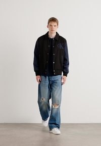 Black and navy bomber jacket with logo on chest, paired with light blue ripped jeans and white sneakers. Model stands against a neutral background.