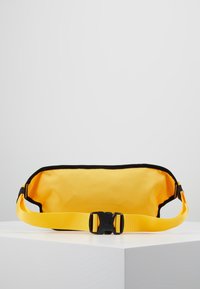 Žlutá látková ledvinka s černým lemem, vybavená nylonovým popruhem a černou plastovou sponou. Hladký povrch a minimalistický design.