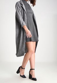 Femme portant une robe courte gris à motifs, un long manteau à carreaux, et des chaussures noires à talons hauts avec bride à la cheville, marchant devant un fond uni.