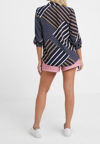 Granatowa bluzka w skośne paski, z podwiniętymi rękawami i geometrycznymi wzorami, zestawiona z jasnoróżowymi szortami i białymi trampkami.