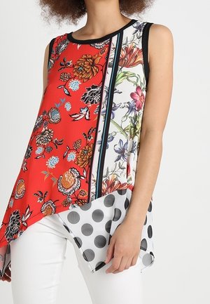 Sleeveless top with asymmetrical hem, featuring red floral, white floral, and black polka dot panels worn with white pants.