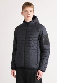 Casaco puffer preto com capuz, apresentando textura acolchoada, fecho frontal e um bolso com fecho no braço esquerdo. Branding minimal visível.