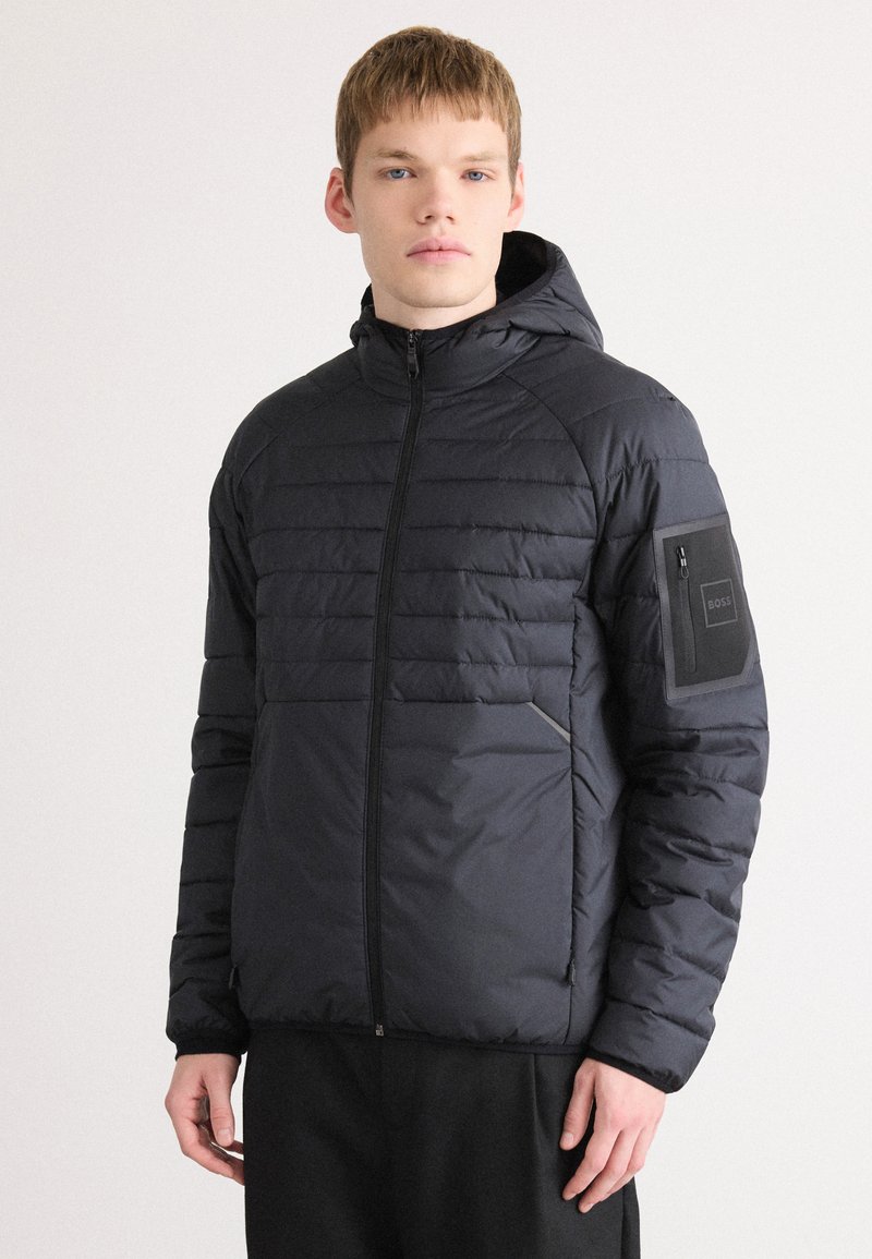 Casaco puffer preto com capuz, apresentando textura acolchoada, fecho frontal e um bolso com fecho no braço esquerdo. Branding minimal visível.
