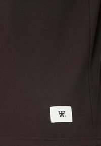 Tamno smeđi teksturirani materijal s malom bijelom etiketom koja sadrži crni logo. Materijal ima uzorak mreže, stvarajući strukturirani izgled.