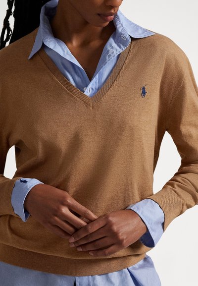 Καφέ πουλόβερ με ντεκολτέ σε σχήμα V από απαλό ύφασμα, φορεμένο πάνω από ένα ανοιχτό μπλε πουκάμισο με γιακά. Διαθέτει ένα μικρό μπλε λογότυπο στο αριστερό στήθος.