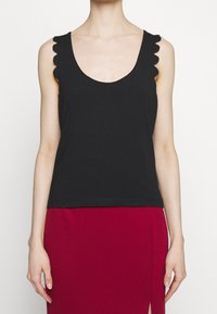 Personne portant un haut noir sans manches avec des emmanchures festonnées et une jupe rouge avec une fente à l'avant, montrée des épaules aux genoux.
