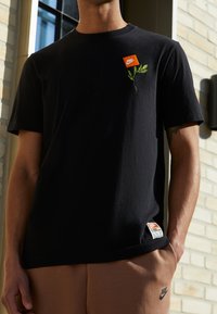 Svart bomulls t-shirt med en liten orange Nike-logotyp och grön växtbrodyr på vänster bröst; etikett synlig på den nedre fållen.