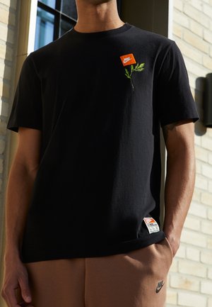 Zwarte katoenen t-shirt met een klein oranje Nike-logo en groene plantenborduursel op de linkerkant van de borst; label zichtbaar op de onderste zoom.
