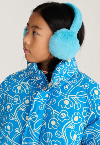 Jeune fille portant une veste fleurie bleu vif et des cache-oreilles bleus assortis et duveteux, regardant sur le côté sur un fond uni.
