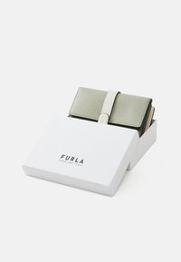 Portefeuille en cuir vert avec une finition texturée, fermé par une sangle blanche, placé à l'intérieur d'une boîte blanche avec le logo "FURLA".