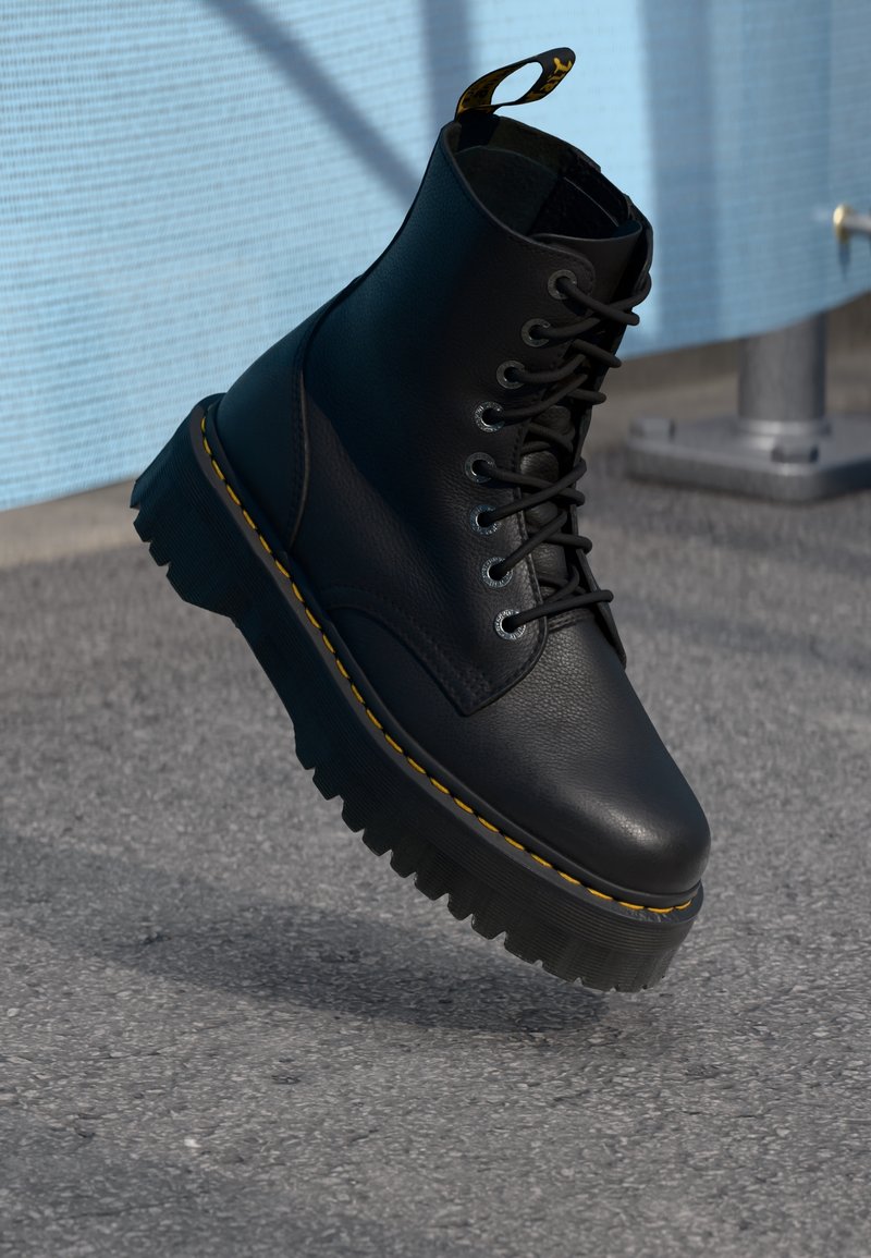 Botte en cuir noir à lacets avec semelle épaisse et crantée et coutures jaunes, suspendue au-dessus d'un sol asphalté près d'un poteau métallique.