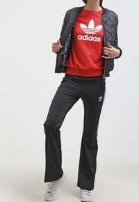 Svart quiltad bomberjacka över en röd Adidas sweatshirt med vit logotyp, svarta byxor med vita ränder och grå sneakers.