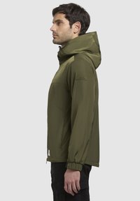 Hellgrüne, leichte Jacke mit Kapuze, elastischen Bündchen und einer mit Reißverschluss versehenen Tasche. Mit einem Logo-Patch auf der linken Schulter.