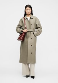 Γυναίκα με μπεζ μακρύ παλτό τύπου τρανς με κουμπιά και ζώνη, εκρού παντελόνι με φαρδιά πόδια, μαύρα μυτερά παπούτσια και σκούρο κόκκινο τσάντα ώμου σε απλό φόντο.