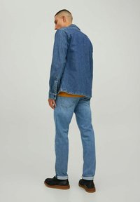 Chaqueta de mezclilla en azul oscuro con cuello clásico y bolsillos frontales, combinada con jeans azul claro y zapatos negros con suela marrón.
