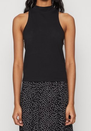 Kobieta ubrana w bezrękawnik z czarnej bluzki z imitacją kołnierzyka oraz czarną spódnicę w białe kropki na jednolitym tle.