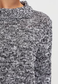 Maglione lavorato a maglia in bianco e nero, con un colletto alto spesso, un motivo testurizzato e una vestibilità ampia. Dettaglio accentuato in rosso visibile.