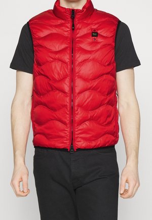 Homme portant une veste matelassée rouge sans manches sur une chemise noire à manches courtes et un pantalon noir, la veste zippée à l'avant avec un petit écusson noir sur la poitrine.