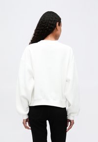 Valkoinen, ylisuuri huppari, jossa on pyöreä kaula-aukko, lasketut olkapäät ja ribattuja hihoja. Sileä pinta, suunniteltu rentoon istuvuuteen.