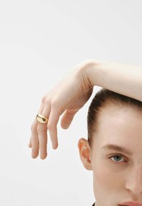 Anillo dorado con un diseño liso y redondeado, llevado en el dedo índice de una mano con piel clara, sobre un fondo blanco liso.