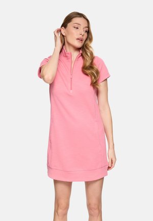 Mujer con un vestido corto de manga corta rosa con cremallera frontal, de pie con una mano tocándose el cabello y mirando ligeramente hacia un lado.