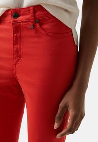 Rote Jeans aus Baumwollmischung mit glatter Textur, ausgestattet mit einem vorderen Knopfverschluss, Seitentaschen und einem kleinen metallischen Logo-Akzent.